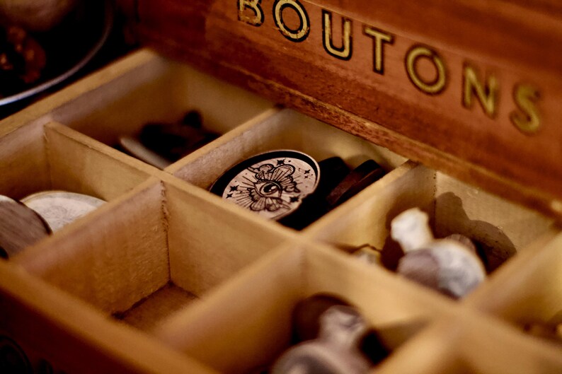 May include: A wooden box with the word "BUTTONS" on the lid. The box is open and contains various buttons, including a round button with a black and white eye design.