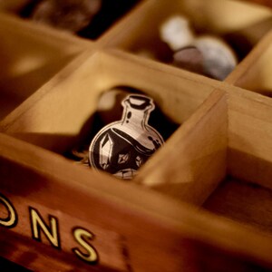May include: A wooden box with compartments, each containing a small, black and white illustration of a potion bottle. The box is labeled "TONS" in yellow letters.