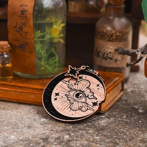 May include: A wooden charm with a black and white design of an eye with a lightning bolt and clouds. The eye is surrounded by stars and a crescent moon.