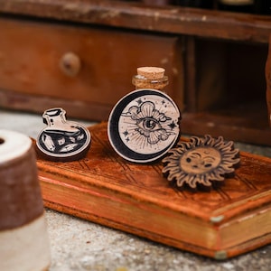 May include: Three wooden pinback buttons with black and white illustrations. The first button features a potion bottle with crystals inside. The second button features a crescent moon with a cloud and an eye. The third button features a sun with a face.