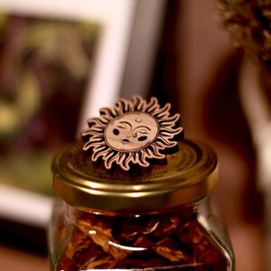 May include: A wooden sun with a face carved into it sits on top of a glass jar filled with dried fruit. The jar has a gold lid.
