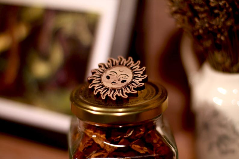 May include: A wooden sun with a face carved into it sits on top of a glass jar filled with dried fruit. The jar has a gold lid.