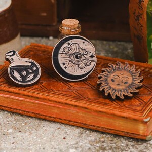 May include: Three wooden pins with a mystical theme. The pins feature a potion bottle, a sun with a face, and a crescent moon with an eye in the center.