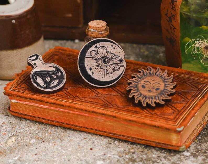 May include: Three wooden pins with a mystical theme. The pins feature a potion bottle, a sun with a face, and a crescent moon with an eye in the center.