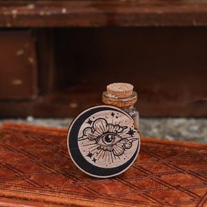May include: A wooden circle with a black background and a white design of an eye with a crescent moon and stars. The circle is sitting on a brown leather-bound book.