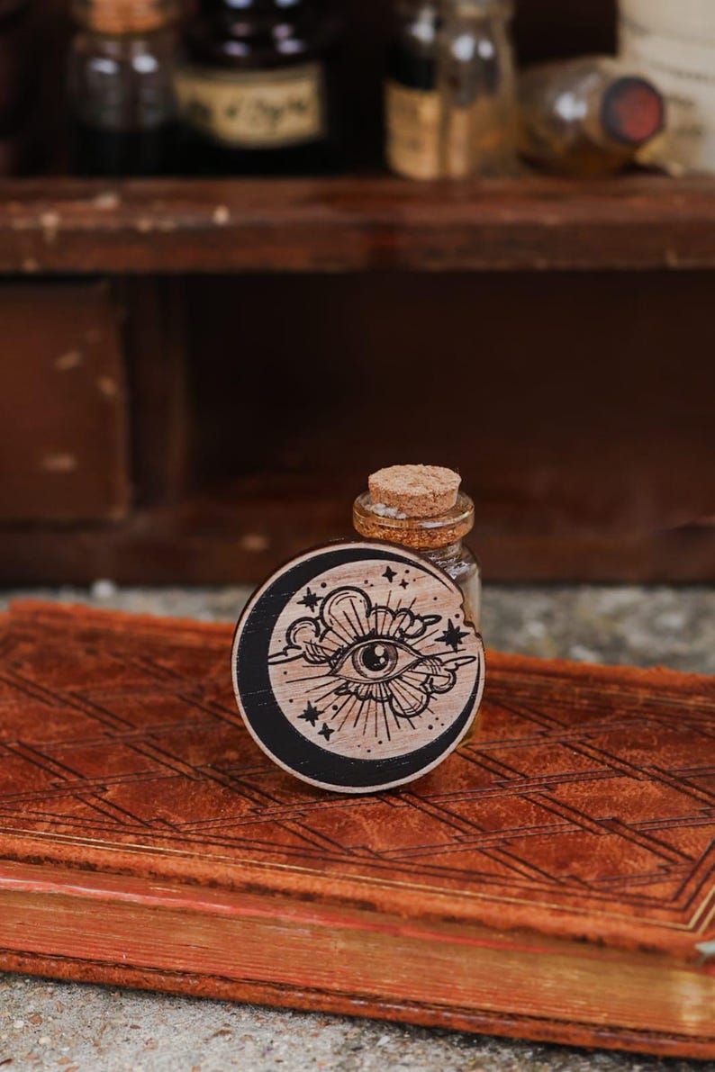 May include: A wooden circle with a black background and a white design of an eye with a crescent moon and stars. The circle is sitting on a brown leather-bound book.