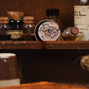 May include: A wooden round stamp with a black and white illustration of an eye with a cloud and stars. The stamp is on a shelf with several glass bottles.