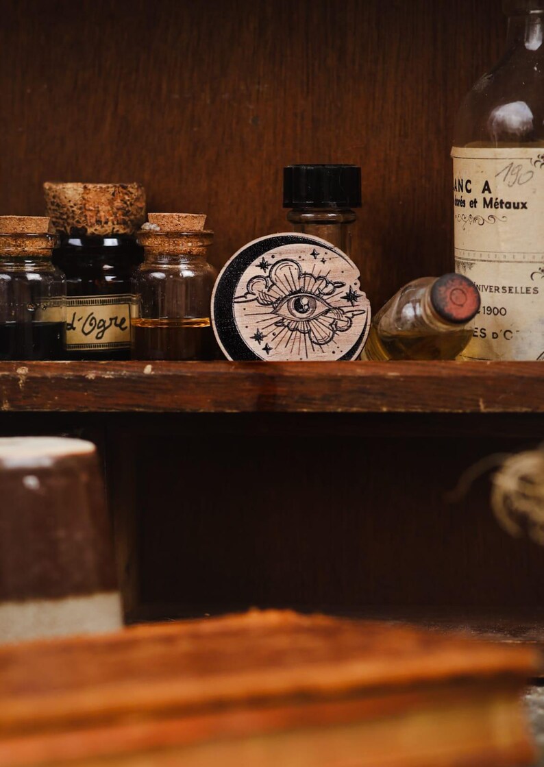 May include: A wooden round stamp with a black and white illustration of an eye with a cloud and stars. The stamp is on a shelf with several glass bottles.
