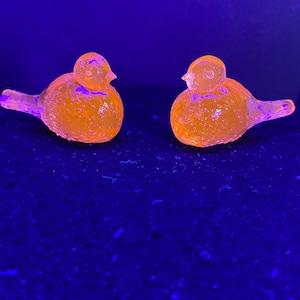 May include: Two orange glass bird figurines with textured surfaces. The birds are illuminated with a bright light against a dark blue background. The birds are facing each other.