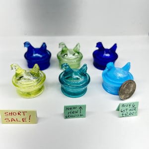 May include: A collection of six colorful glass hen-shaped containers in shades of blue, green, and yellow. The containers are displayed on a white surface with small signs that read "SHORT SALE!" and "BUY 6 GET ONE FREE!"