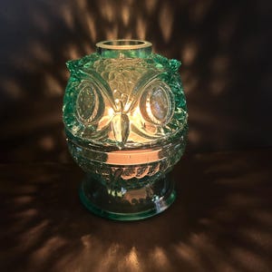 May include: A teal-colored glass owl-shaped candle holder. The candle holder has a detailed owl face design with large eyes and a textured surface. The candle inside casts a warm glow, creating a cozy ambiance.