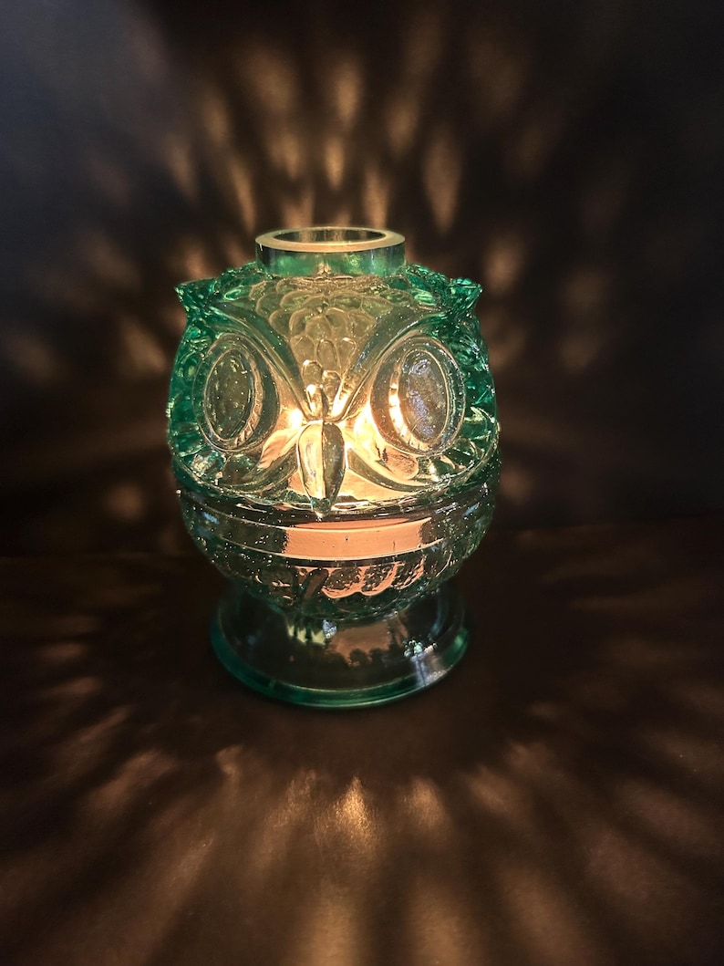 May include: A teal-colored glass owl-shaped candle holder. The candle holder has a detailed owl face design with large eyes and a textured surface. The candle inside casts a warm glow, creating a cozy ambiance.