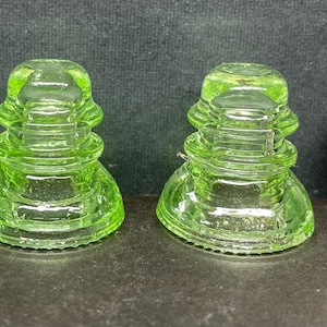 May include: Three small, translucent lime green glass insulators with a tiered, stepped design. Each piece features a wide circular base and a narrower top section. A United States quarter coin is placed to the right for scale comparison.