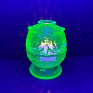 May include: A green glass owl-shaped decorative item with a pedestal base. The owl has large, round eyes and a textured surface. The item glows brightly against a dark blue background.