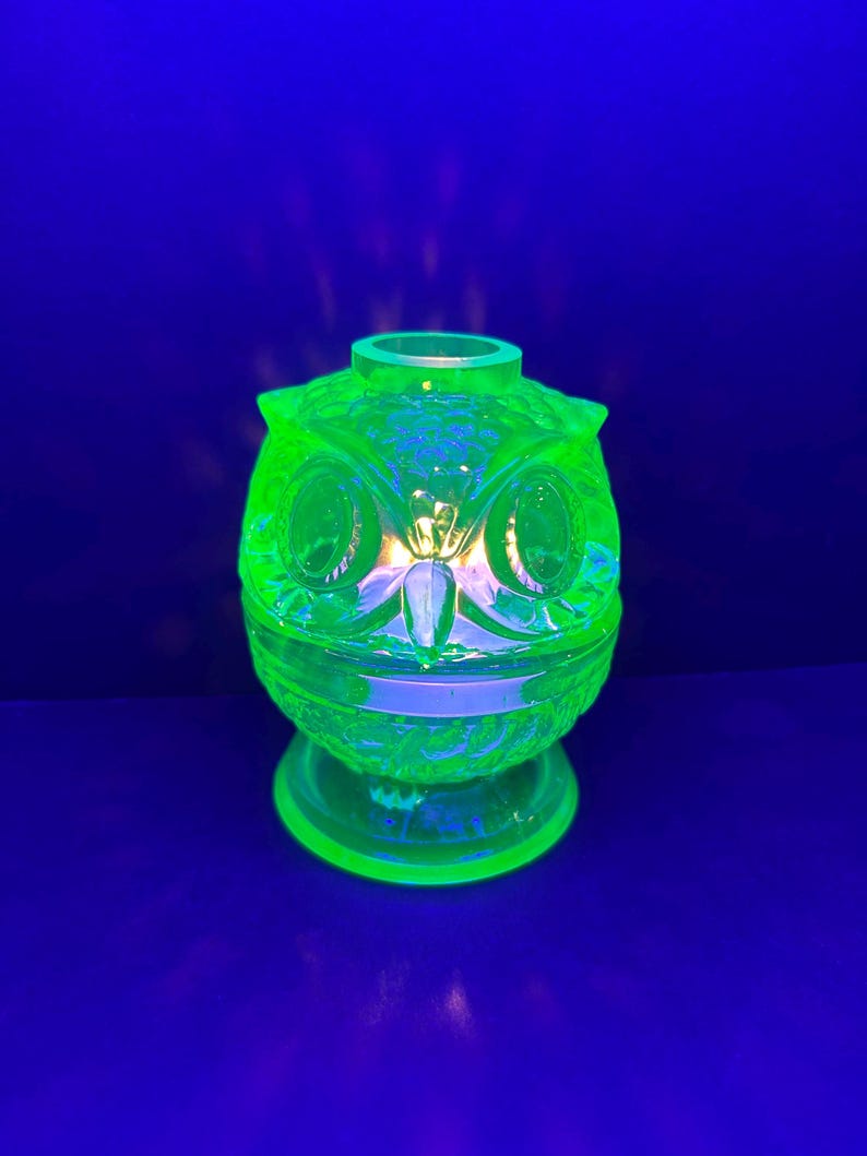 May include: A green glass owl-shaped decorative item with a pedestal base. The owl has large, round eyes and a textured surface. The item glows brightly against a dark blue background.