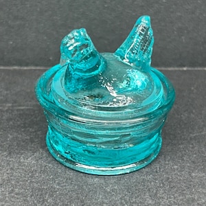 May include: A turquoise glass bottle stopper with a bird-like design. The stopper features a round base and a detailed top with two raised elements resembling wings or ears. The glass has a textured appearance, and the overall design is decorative.