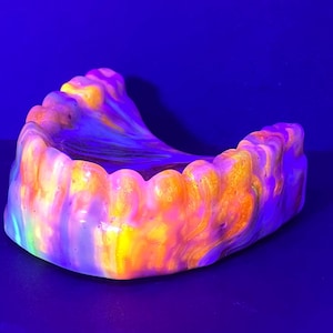 May include: A dental model of a lower jaw featuring a vibrant, swirling marbled pattern of neon orange, yellow, and purple hues. The surface appears smooth and glossy, reflecting light under ultraviolet illumination against a dark background.