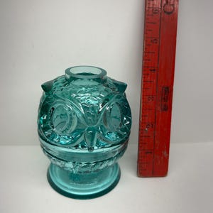 May include: A teal-colored glass owl-shaped container with detailed facial features and a round opening at the top. A vintage red ruler is positioned next to the container, showing measurements in inches.