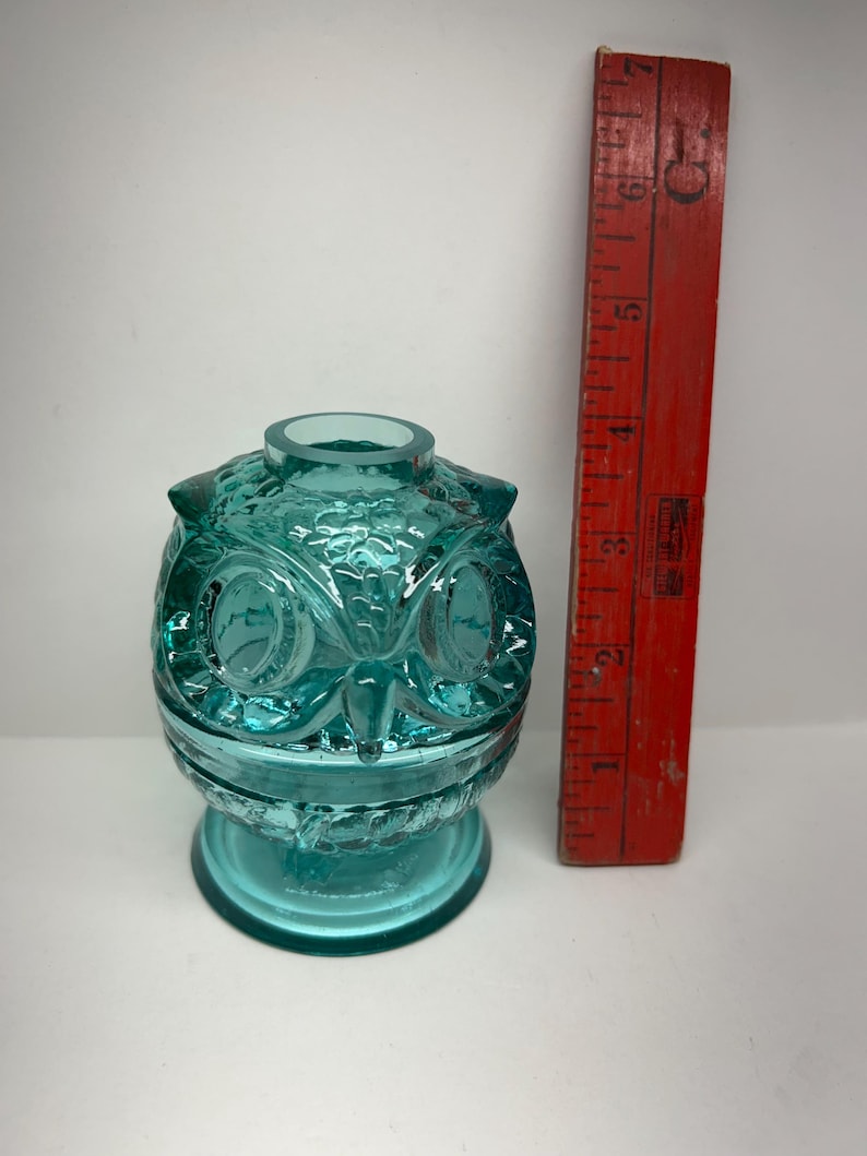 May include: A teal-colored glass owl-shaped container with detailed facial features and a round opening at the top. A vintage red ruler is positioned next to the container, showing measurements in inches.