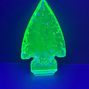 May include: A green glass arrowhead figurine with a textured surface. The arrowhead is illuminated with a blacklight, causing it to glow.
