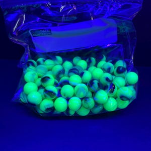 May include: A clear plastic bag filled with glowing green marbles. Each marble has a swirl of blue and black. The bag has a white label with the words "DATE: CONTENTS" printed on it. The background is a dark blue.