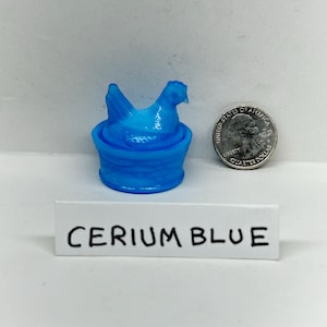 May include: A cerium blue figurine of a hen sitting in a basket. The figurine is next to a U.S. quarter. The text "CERIUM BLUE" is on a white label.