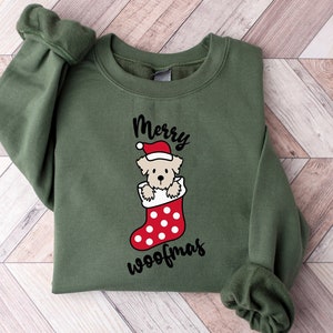 May include: Green Christmas sweatshirt with a white dog wearing a Santa hat peeking out of a red and white stocking with white polka dots. The text "Merry Woofmas" is printed below the stocking.