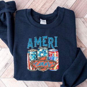 Puede incluir: Una sudadera azul marino con un gráfico de tres latas rojas, blancas y azules con el texto "AMERI can" en un letrero de madera.