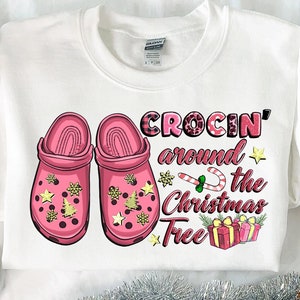 Könnte beinhalten: Weißer Pullover mit einem rosafarbenen und weißen Weihnachtsmotiv. Das Motiv zeigt zwei rosa Clogs mit goldenen Glitzer und den Text "Crocin' around the Christmas Tree".