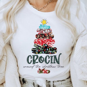 Könnte beinhalten: Weißes T-Shirt mit einem Weihnachtsbaumdesign aus bunten Crocs. Der Text "Crocin around the Christmas tree" ist unter dem Baum gedruckt.