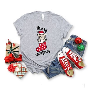 May include: A gray t-shirt with a Christmas-themed design featuring a dog wearing a Santa hat in a red and white stocking. The text "Merry Woofmas" is printed below the dog.