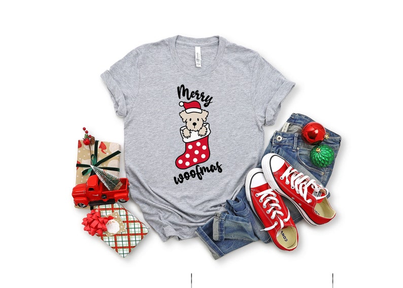 May include: A gray t-shirt with a Christmas-themed design featuring a dog wearing a Santa hat in a red and white stocking. The text "Merry Woofmas" is printed below the dog.