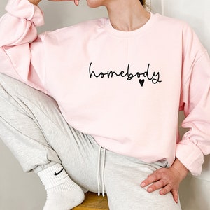 May include: A light pink sweatshirt with the word "homebody" in a handwritten font, with a small black heart. The sweatshirt has long sleeves and is paired with gray sweatpants. The person is wearing white socks.