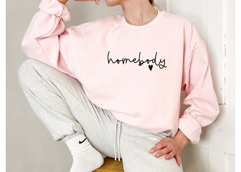 May include: A light pink sweatshirt with the word "homebody" in a handwritten font, with a small black heart. The sweatshirt has long sleeves and is paired with gray sweatpants. The person is wearing white socks.