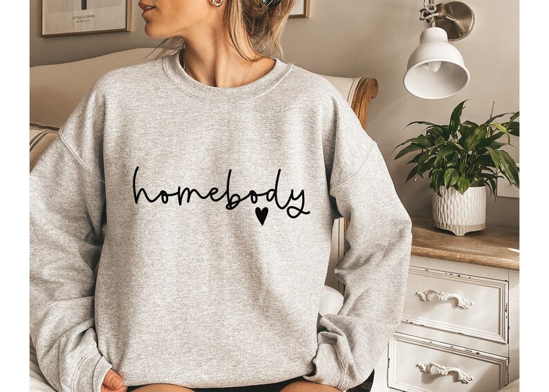 May include: A light gray sweatshirt with the word "homebody" in black script, with a small black heart below the word. The sweatshirt is a crew neck style and appears to be made of a soft material. The overall theme is casual wear.