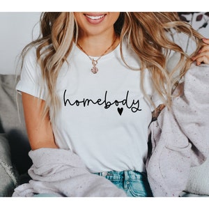 May include: White t-shirt with the word "homebody" in black script and a small black heart. The shirt is paired with blue jeans and a light purple sweater. A gold necklace is visible.