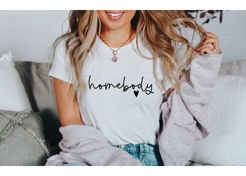 May include: White t-shirt with the word "homebody" in black script and a small black heart. The shirt is paired with blue jeans and a light purple sweater. A gold necklace is visible.