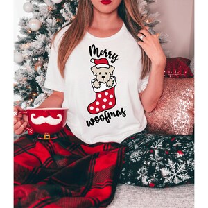 May include: White t-shirt with a Christmas-themed graphic of a dog wearing a Santa hat and peeking out of a red and white stocking. The text "Merry Woofmas" is printed below the graphic.