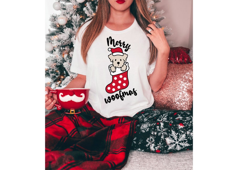 May include: White t-shirt with a Christmas-themed graphic of a dog wearing a Santa hat and peeking out of a red and white stocking. The text "Merry Woofmas" is printed below the graphic.