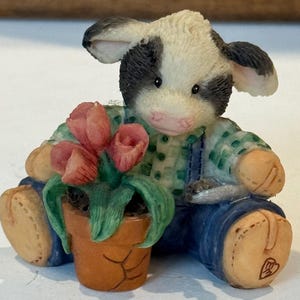 May include: A figurine of a calf wearing a green plaid shirt and blue overalls, holding a terracotta pot with pink tulips. The calf has black and white markings and is sitting down. The figurine has a whimsical, handcrafted appearance.
