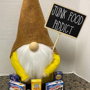 May include: A yellow gnome with a brown hat and a white beard holds a sign that says "Junk Food Addict". The gnome is surrounded by miniature boxes of junk food, including Twinkies, Wheat Thins, Cup Cakes, and Airheads.