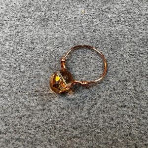 May include: A handmade copper wire ring featuring a faceted, amber-colored crystal bead. The ring's band is a continuous loop of copper wire, with the bead securely wrapped at the top. The ring is set against a gray textured background.