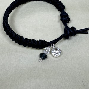 May include: Black braided bracelet with a dangling charm. The charm features a large, clear, round crystal and two smaller beads. The bracelet is knotted for closure. A simple, elegant piece of jewelry.