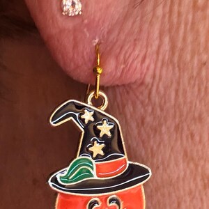 Puede incluir: Un pendiente dorado con temática de Halloween. El pendiente presenta una calabaza con un sombrero de bruja. La calabaza es naranja con una cara negra y el sombrero es negro con estrellas doradas. También se ve un pequeño pendiente de botón redondo y transparente.