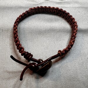 May include: Braided brown leather bracelet with a dark brown button clasp. The bracelet is made of tightly woven leather strands, creating a textured appearance. The clasp is a dark, rounded button with a loop for closure. A simple, rustic accessory.