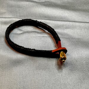 May include: A black braided bracelet with a brown leather accent and a gold-colored clasp. The bracelet is circular and appears to be handmade. The background is a light gray fabric.