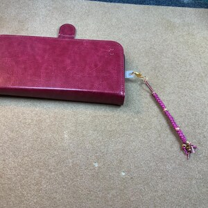 May include: A burgundy leather phone case with a magnetic closure and a matching lanyard. The lanyard is a braided purple cord with gold-colored accents and a small charm.