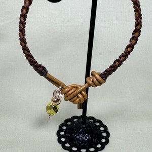 May include: A brown leather necklace with a braided design. The necklace features a knot design and a dangling charm with a yellow-green faceted bead and silver beads. The necklace is displayed on a black metal stand.