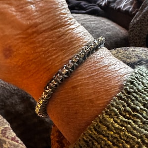 Puede incluir: Una pulsera plateada con piedras claras de corte cuadrado se lleva en la muñeca. La pulsera está hecha de pequeños cuadrados unidos, cada uno con una piedra. La muñeca es visible, con la piel mostrando arrugas y edad.