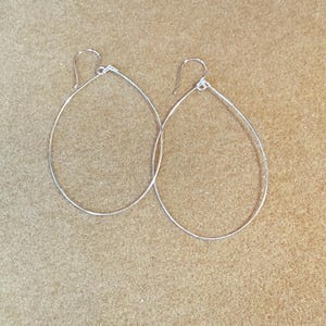 May include: A pair of silver teardrop hoop earrings. The earrings have a simple, minimalist design with a slender, smooth finish. The hooks are visible at the top, ready for wear. The earrings are displayed against a neutral background.